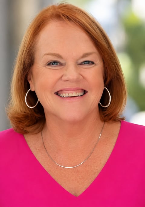 Smiling woman with shoulder-length reddish-brown hair wearing a bright pink top and silver jewelry